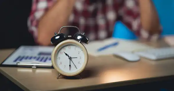 Despertador sobre un escritorio con informes y un trabajador preocupado detrás