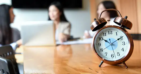 Despertador sobre la mesa de una oficina con trabajadores al fondo